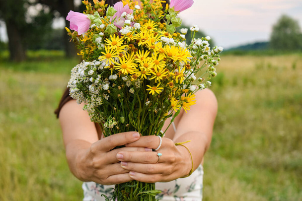 Focus shifts bouquet multiracial young woman stands picturesque meadow at twilight. Face remains hidden behind bouquet wildflowers, drawing attention to vibrant colors and enchanting atmosphere scene - Photo, Image