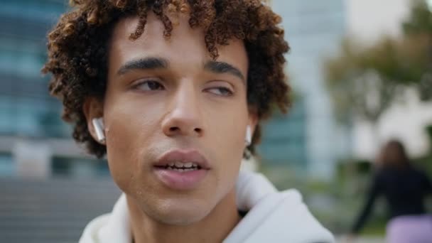 Mladý muž mluví bezdrátovými sluchátky na portrétu v centru. Curly teenager tvář dělat hovor mluvící sluchátka venku. Klidný hipster používající modrozubé airpody komunikující na ulici. Technologický koncept - Záběry, video