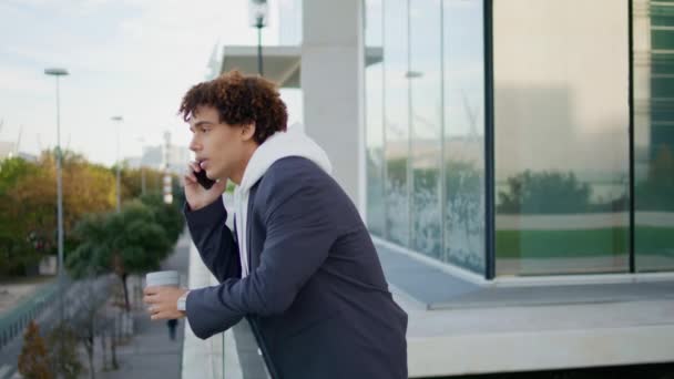 Κέρλι hipster καλώντας smartphone στο γυάλινο μπαλκόνι από κοντά. Κομψός μαθητής που κρατάει φλυτζάνι τσαγιού βλέποντας θέα στην πόλη. Χαμογελώντας έφηβος έχει ευχάριστη συνομιλία στο κινητό τηλέφωνο. Μοντέρνος τύπος που χρησιμοποιεί τηλέφωνο μόνος - Πλάνα, βίντεο