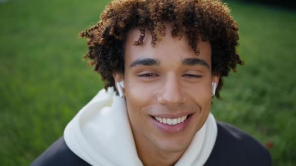 Šťastnej chlap, co používá sluchátka na zeleným trávníkovým portrétu. Student sluchátek se zubatě usmívá a vychutnává hudbu v přírodě. Mladý muž hledící kamerou zblízka do parku. Latinky teen tvář zírá objektiv pocit radosti - Záběry, video