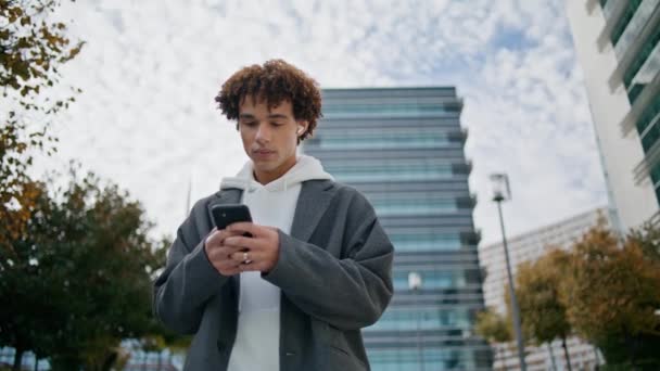 Krullende tiener typt smartphone op straat close-up. Jongeman luistert muziek met behulp van oortjes buiten. Knappe hipster zoekt online met mobiel in de herfst. Gerichte man scrollen telefoon scherm - Video