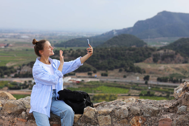 Ελκυστική νεαρή ταξιδιωτική blogger με σακίδιο στέκεται στο λόφο μετά από πεζοπορία μέχρι κατέχει smartphone, επικοινωνεί με τους οπαδούς της και δείχνει το τοπίο στα μέσα κοινωνικής δικτύωσης. Ατομικό ταξίδι ασφαλείας στην Ευρώπη. - Φωτογραφία, εικόνα