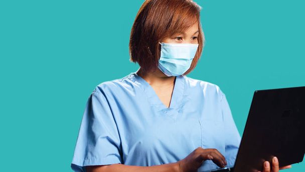 Közelkép mosolygós boldog egészségügyi szakember segítségével laptop keresni orvosi információk online. Ázsiai nővér visel professzionális klinikai védő egyenruhát, izolált stúdió háttér - Fotó, kép