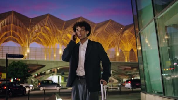 Young man talking phone at illuminated city. Smiling businessman calling smartphone waiting in airport bus stop. Curly entrepreneur waving arm meeting relatives in downtown district. Business trip - Footage, Video