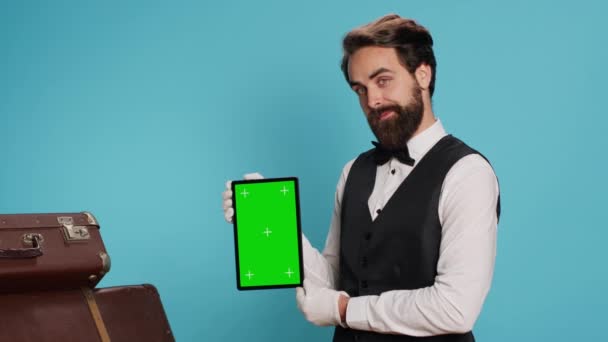 Doorkeeper holds tablet with isolated display, posing against blue background with formal uniform. Professional hotel concierge employee showcasing greenscreen copyspace on gadget. - Footage, Video