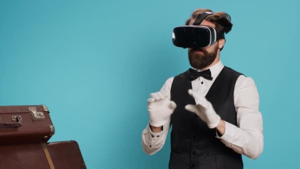 Ο σύγχρονος γκρουμ δοκιμάζει γυαλιά VR σε μπλε φόντο, χρησιμοποιώντας τεχνολογία εικονικής πραγματικότητας στην κάμερα. Εκλεπτυσμένος θυρωρός στέκεται δίπλα στο σωρό αποσκευών, τουριστική φιλοξενία έννοια της βιομηχανίας. - Πλάνα, βίντεο