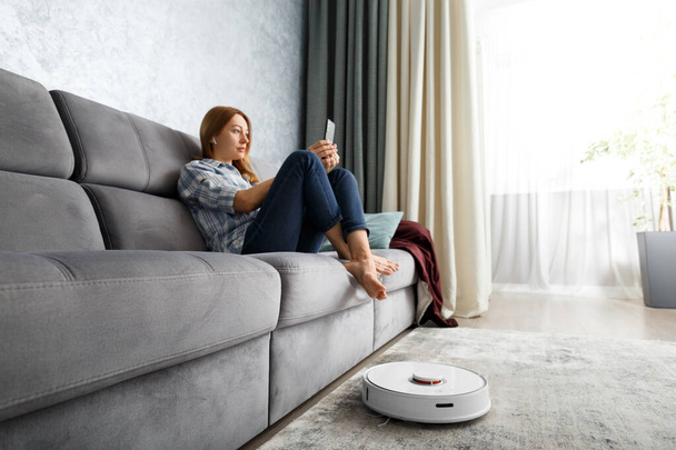 Робот пылесос в интерьере гостиной. Девушка сидит на диване и управляет умным пылесосом с помощью смартфона. Концепция умного дома, технологии, домашняя работа. - Фото, изображение