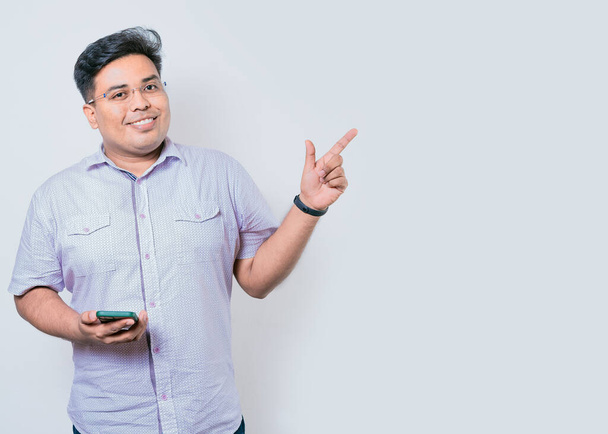 Young latin man holding cellphone pointing to side isolated. Happy guy holding cellphone pointing to the side isolated - Photo, Image