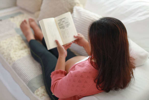 Смотрите на счастливую молодую женщину, которая сидит и отдыхает на диване в гостиной, читая книгу с релаксацией. Ожидаемая мать готовится к родам во время беременности - Фото, изображение