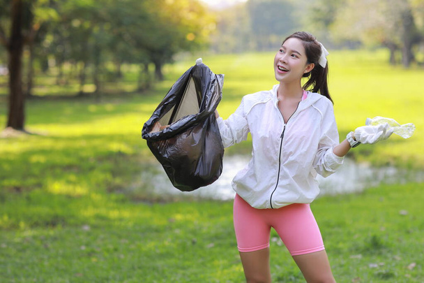 Na de ochtend lopen jonge mooie Aziatische vrijwilliger vrouw houden vuilniszak en het ophalen van afval van het park gazon en tonen plastic fles en volledige zwarte tas tijdens een vrijwilliger milieu opruimen  - Foto, afbeelding