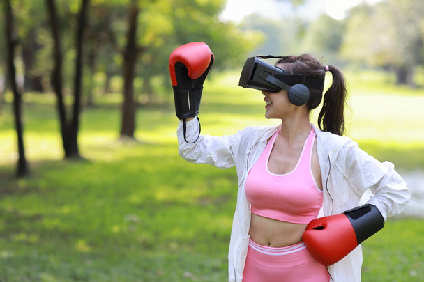 Счастливая азиатка фитнес-спортсменка с технологией бокса виртуальной реальности виртуальной реальности, занимающаяся аэробной тренировкой для боксёрского пунша, поднимая руки под деревом в парке - Фото, изображение