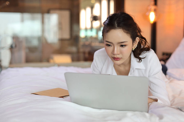 Aktiivinen aasialainen nainen rento makaa sängyllä ja työskentelee tietokoneella kannettavan hotellin, kauneus liikenainen makuuhuone tausta. Elämäntapateknologian käsite - Valokuva, kuva