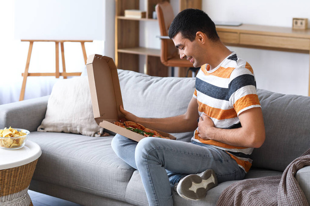 Νεαρός άνδρας με κουτί νόστιμη πίτσα κάθεται στον καναπέ στο σπίτι - Φωτογραφία, εικόνα