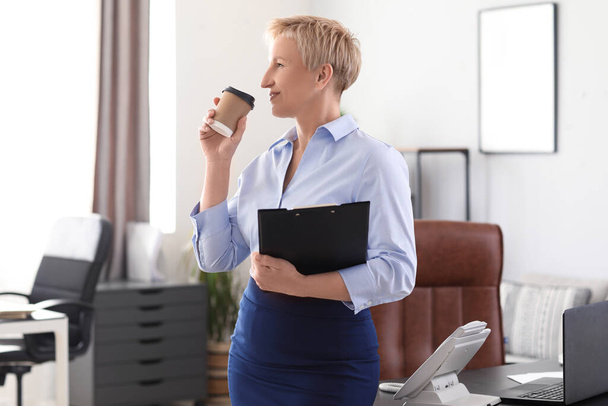 Rijpe zakenvrouw met documenten en koffie in kantoor - Foto, afbeelding