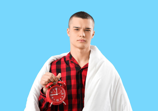 Displeased young man in pajamas, with blanket and alarm clock on blue background - Photo, Image