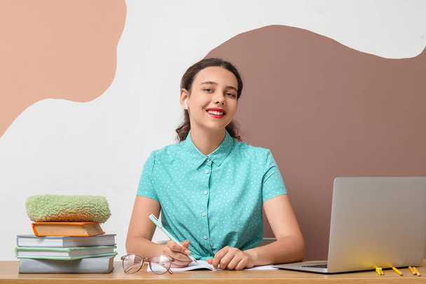 Estudante feminina em fones de ouvido com laptop fazendo lição de casa na mesa perto da parede de cor - Foto, Imagem