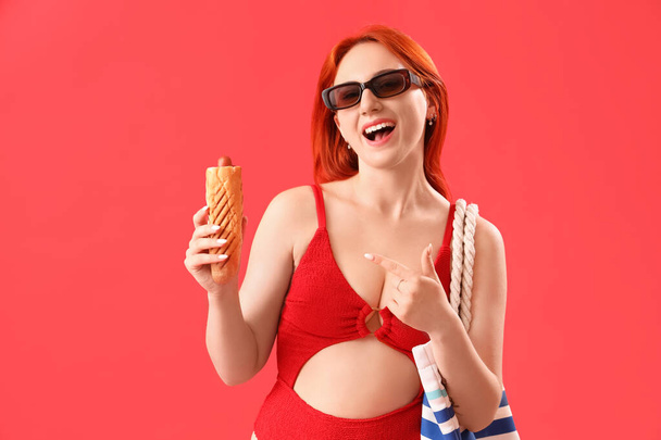 Bella giovane donna in costume da bagno, con borsa da spiaggia e hot dog francese su sfondo rosso - Foto, immagini