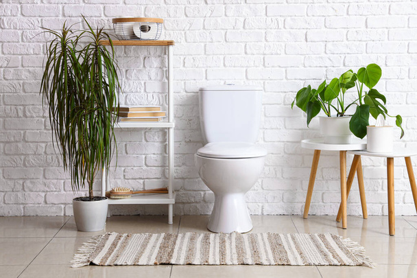 Интерьер светлого туалета с керамической унитазом, полки и комнатные растения рядом с белой кирпичной стеной - Фото, изображение