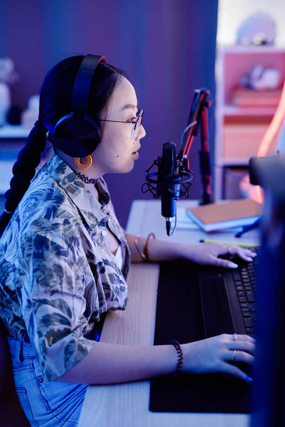 Vista lateral de la adolescente jugando videojuego en PC y hablando con el micrófono mientras se transmite en vivo en luz de neón azul - Foto, imagen