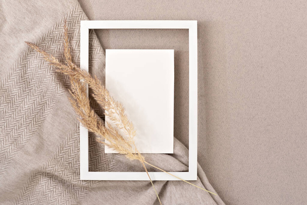 Elegant neutral beige greeting postcard or wedding invitation design template. Blank paper card mock up, empty picture frame border, dried meadow grass on a crumpled knitted fabric background - Photo, Image
