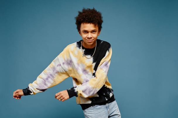 Minimal portrait of black teenage boy wearing tie dye shirt and dancing over blue background, copy space - Photo, Image