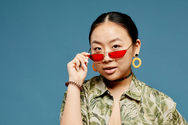 Minimal portrait of Asian teenage girl wearing trendy colored sunglasses over blue background, copy space - Foto, Imagen