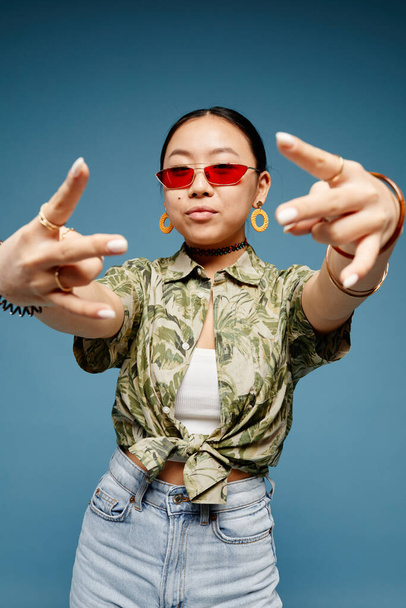 Vertical portrait of trendy teenage girl wearing colored sunglasses and gesturing over blue background - Photo, Image