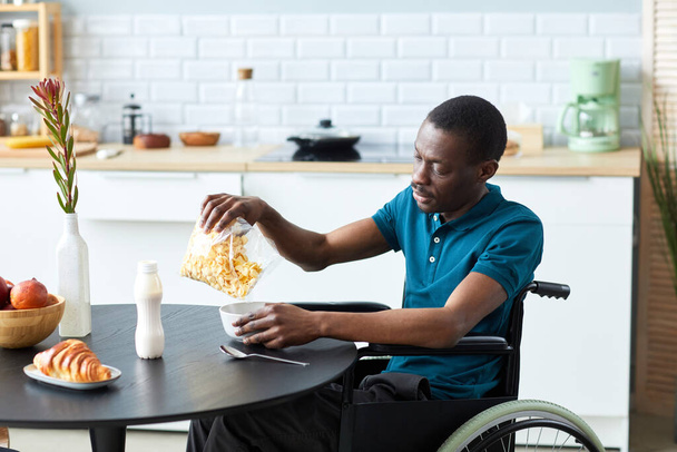 Вид сбоку портрет черного взрослого мужчины с инвалидностью, кушающего хлопья на завтрак в элегантной домашней кухне, пространство для копирования - Фото, изображение