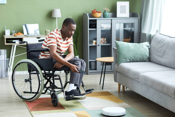 Портрет сучасної чорношкірої людини з інвалідністю за допомогою бездротового робота-пилососа в домашньому інтер'єрі, копіювання простору - Фото, зображення