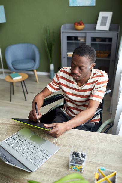 Retrato vertical do homem afro-americano em cadeira de rodas usando caneta tablet enquanto trabalhava com computador em casa - Foto, Imagem