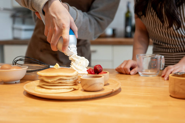 Zugeschnittenes Bild eines Mannes, der Schlagsahne auf Pfannkuchen sprüht, während er Pfannkuchen mit seiner Freundin in der Küche kocht. - Foto, Bild