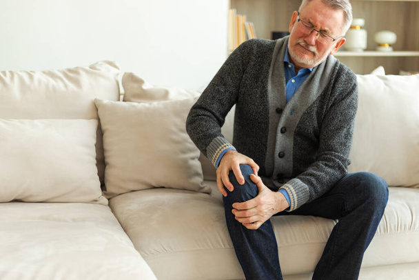 Knee pain arthritis body sick health care concept. Unhappy middle aged senior man suffering from knee ache sitting on sofa at home. Mature old senior grandfather touching leg feeling pain hurt in knee - Photo, Image