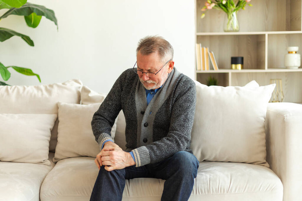 Knee pain arthritis body sick health care concept. Unhappy middle aged senior man suffering from knee ache sitting on sofa at home. Mature old senior grandfather touching leg feeling pain hurt in knee - Photo, Image