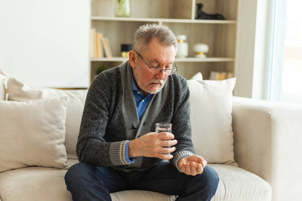 Middle aged senior man holding medical pill and glass of water. Mature old senior grandfather taking medication cure pills vitamin. Age prescription medicine healthcare therapy concept - Photo, Image