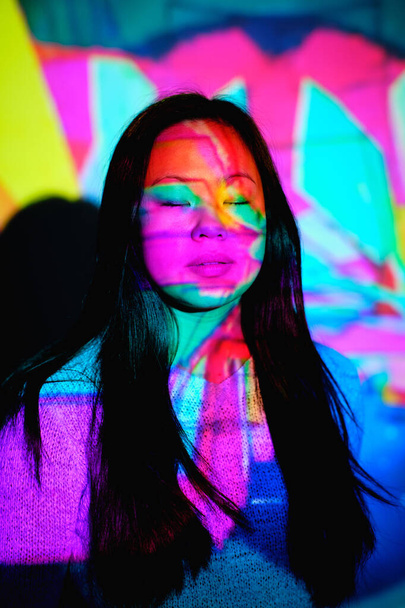 Klidná asijská fena s dlouhými černými vlasy stojící se zavřenýma očima u stěny pod jasným vícebarevným neonovým osvětlením ve stylové místnosti - Fotografie, Obrázek