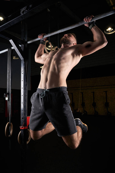 Full body of muscular shirtless male athlete doing pull ups on bar during intense training in dark gym - Photo, Image