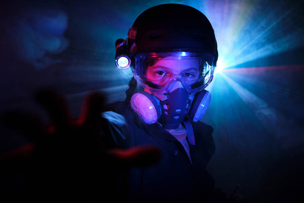 Niño serio en máscara protectora de gas mirando a la cámara con el brazo extendido mientras roba menta oscura con luces de neón brillantes - Foto, imagen