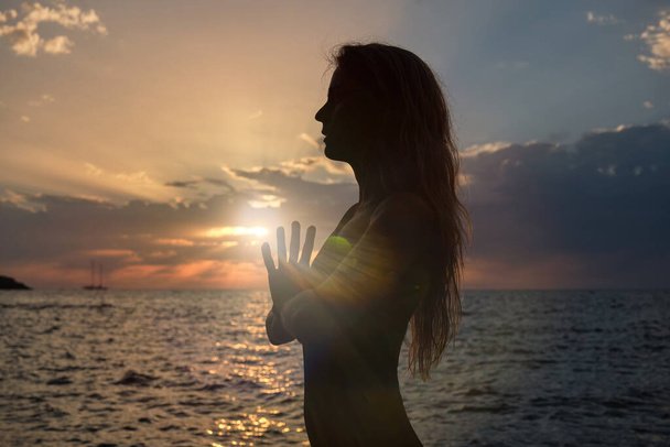 Vista lateral de silueta de mujer anónima meditando con las manos apretadas cerca del mar ondulante durante el atardecer mientras está de pie en Tadasana y haciendo yoga - Foto, Imagen