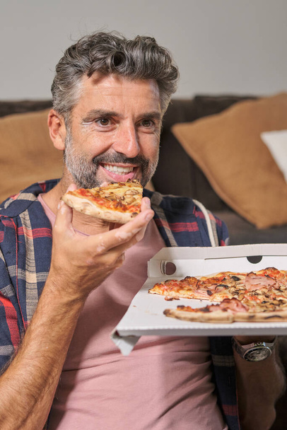 Positieve bebaarde man draagt casual kleding eten heerlijke pizza en weg te kijken terwijl zitten thuis - Foto, afbeelding