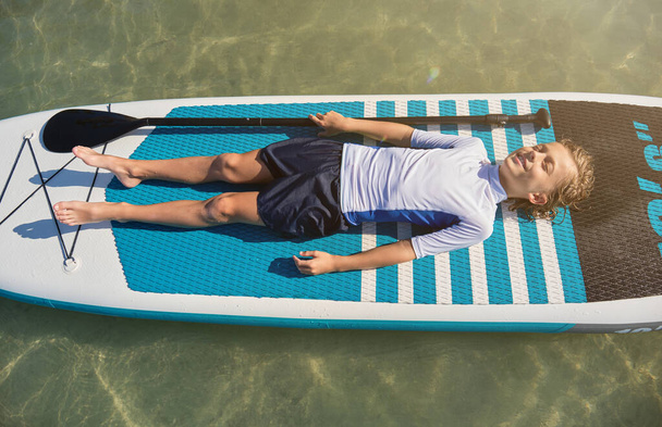 Od shora celé tělo šťastný chlapec v aktivním oblečení ležící na paddleboardu a relaxaci uprostřed moře během letních prázdnin - Fotografie, Obrázek