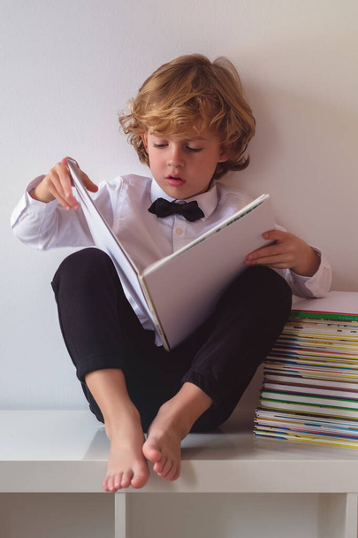 Целое тело очаровательного босоногого мальчика в формальной одежде читает интересную книгу, сидя на белом фоне в светлой комнате - Фото, изображение
