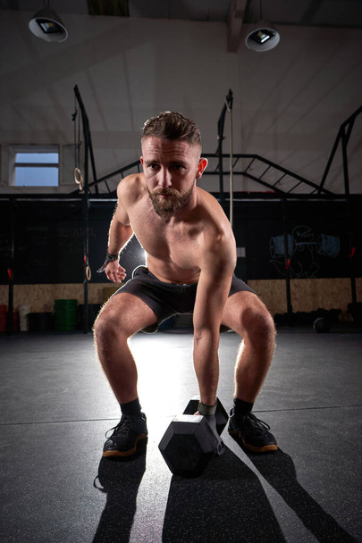 Full length of shirtless sportsman doing squat exagon dumbbell during intense λειτουργική προπόνηση στο γυμναστήριο στο αμυδρό φως - Φωτογραφία, εικόνα