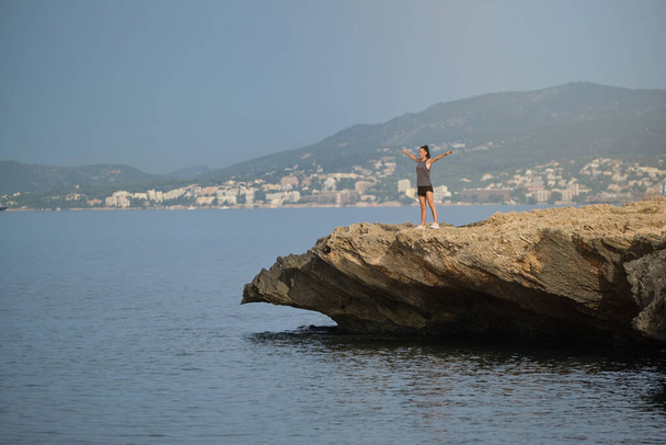 Jovem fêmea de sportswear em pé em penhasco rochoso cercado por mar ondulante contra cidade montanhosa e desfrutando de liberdade com braços estendidos - Foto, Imagem