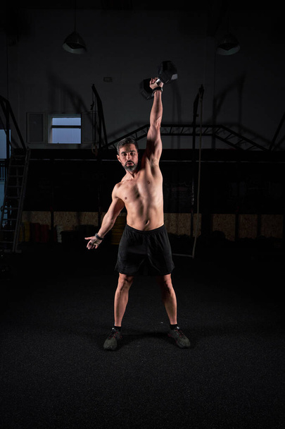 Plná délka sportovního sportovce s nahým trupem v krátkém zvedání těžké šestiúhelník činka při tréninku v tmavé tělocvičně - Fotografie, Obrázek