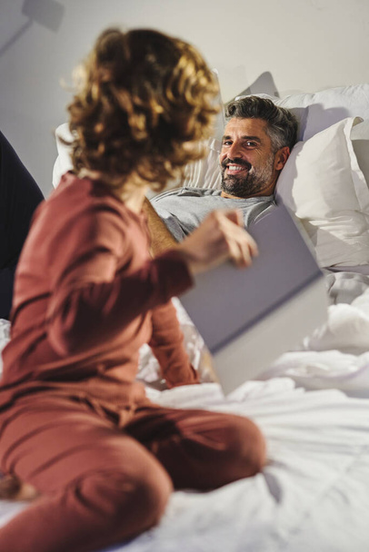 Homme souriant couché sur le lit et regardant garçon avec le livre tout en parlant les uns avec les autres dans une chambre confortable à la maison - Photo, image