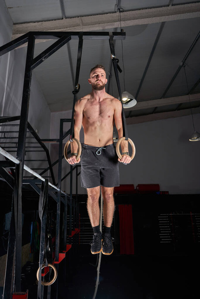 Полное тело мускулистого спортсмена с обнаженным туловищем, висящим на гимнастических кольцах во время функциональной тренировки в тусклом свете - Фото, изображение