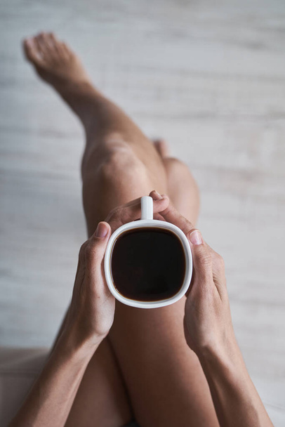 Bovenaanzicht van anonieme vrouw zittend met gekruiste benen en met een mok hete aromatische americano tijdens het ontbijt in de ochtend thuis - Foto, afbeelding