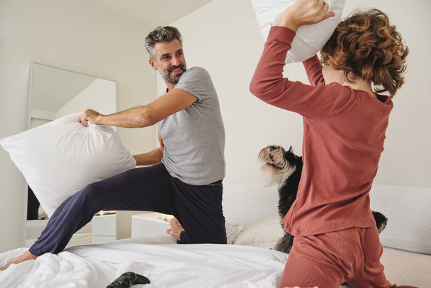 Low angle of mature bearded father and curly child looking at each other while fighting with pillows and playing with dog - Photo, Image