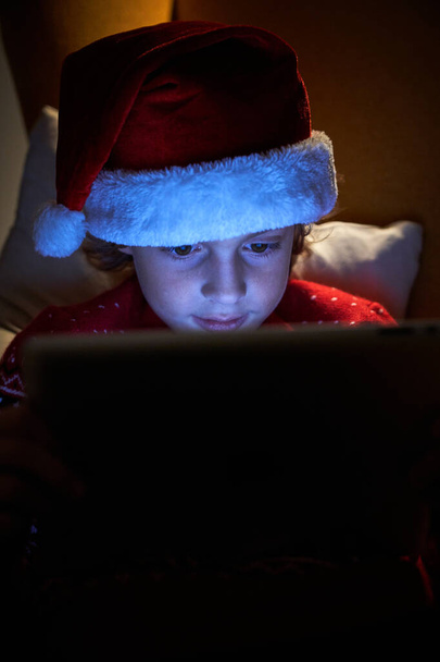 Curieux petit garçon dans le chapeau de Père Noël en utilisant une tablette moderne tout en étant assis dans un fauteuil confortable à la maison le soir - Photo, image