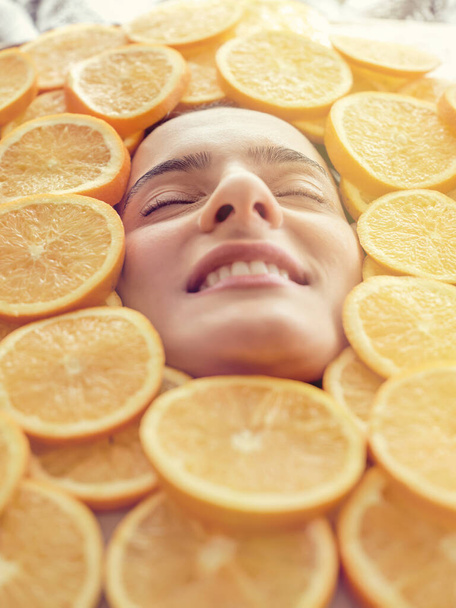 Gezicht van positieve vrouw liggend in grote hoop sappige sinaasappels met gesloten ogen - Foto, afbeelding